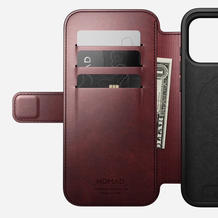The Nomad Modern Leather Folio for iPhone 17 Pro Max in Burgundy Horween leather is shown open, featuring three card slots with two cards and a folded US dollar bill, plus a magnetic closure flap and partial phone compartment view.