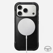 A Nomad Rugged Leather Case for iPhone 17 Pro in black, made from Horween leather, is shown face down with three camera lenses and a circular MagSafe-compatible charger attached to the back.