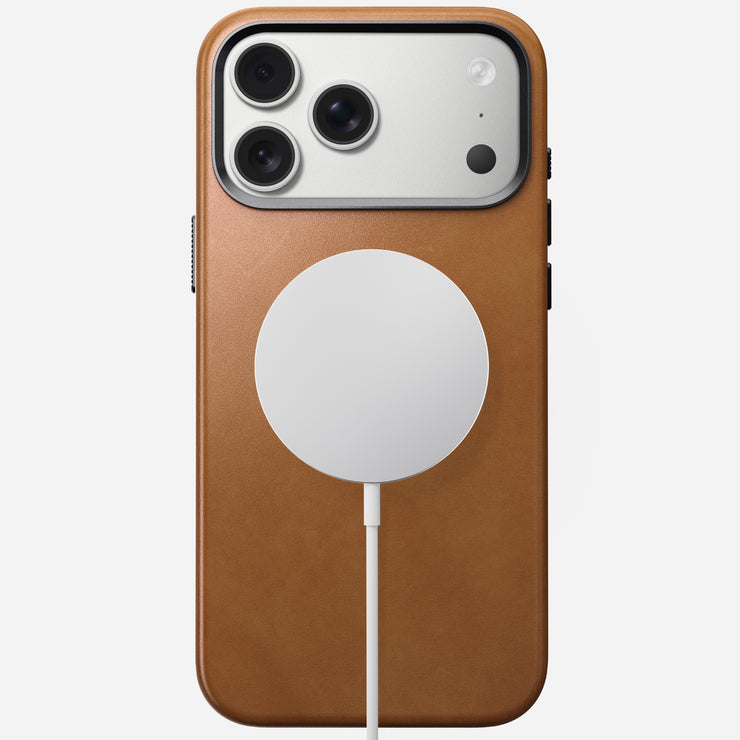 The Nomad Traditional Leather Case in English Tan is shown on an iPhone 17 Pro Max, featuring MagSafe compatibility, three rear cameras, and a charging disc attached to the center of the sustainable leather case with a white cable.