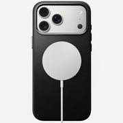 A Nomad Traditional Leather Case in black, made from full-grain leather for iPhone 17 Pro Max, is shown face down with a triple-lens camera and a white circular MagSafe wireless charger attached to its back.