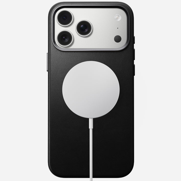 A Nomad Traditional Leather Case in black, made from full-grain leather for iPhone 17 Pro Max, is shown face down with a triple-lens camera and a white circular MagSafe wireless charger attached to its back.