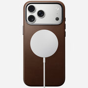 An iPhone 17 Pro Max in a Nomad Traditional Leather Case (brown) lies face down with a circular MagSafe charger attached by a white cable, showing three rear cameras and a camera control button.