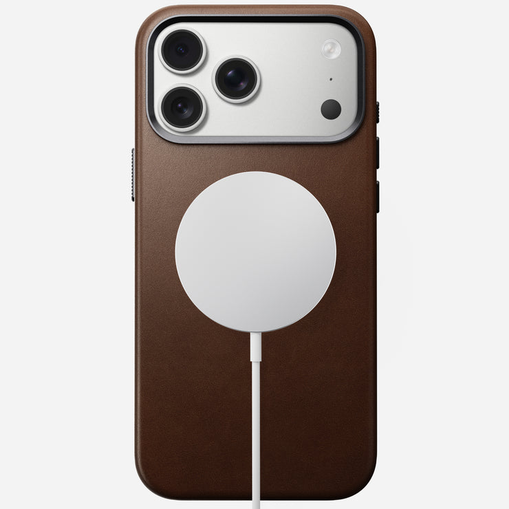 An iPhone 17 Pro Max in a Nomad Traditional Leather Case (brown) lies face down with a circular MagSafe charger attached by a white cable, showing three rear cameras and a camera control button.