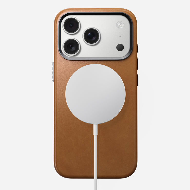 An iPhone 17 Pro in a Nomad Traditional Leather Case (English Tan - C) features three rear cameras and a flash at the top left, with a white MagSafe-compatible wireless charger attached and centered on its sustainably sourced leather back.