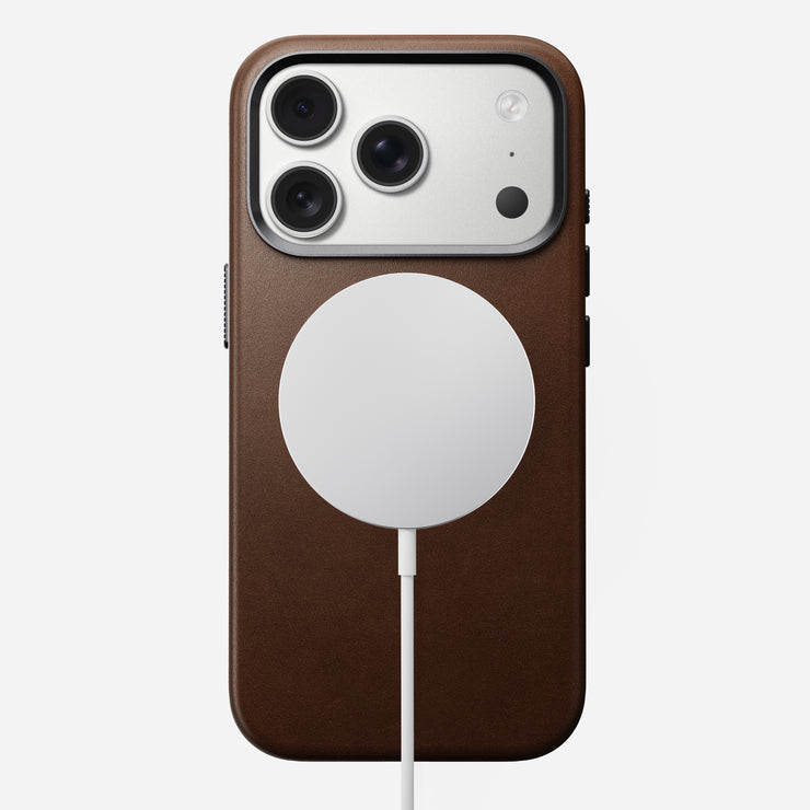 An iPhone 17 Pro in a Nomad Traditional Leather Case (Brown) with MagSafe compatibility, featuring a circular wireless charger attached near the camera lenses.