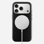 The Nomad Modern Case - iPhone 17 Pro in Vulcan Black, featuring triple rear cameras and TPU drop protection, rests on a circular white wireless charging pad connected by cable against a plain white background.