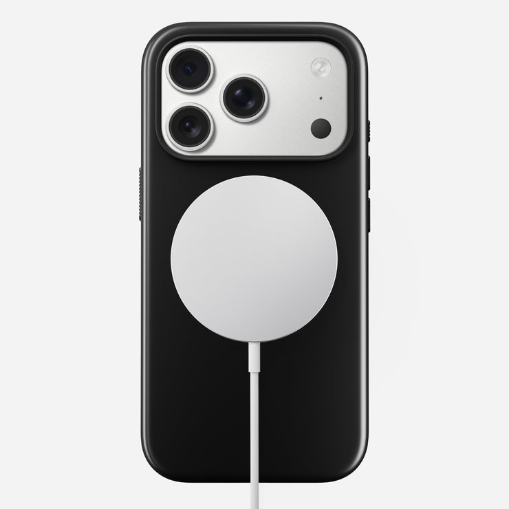 The Nomad Modern Case - iPhone 17 Pro in Vulcan Black, featuring triple rear cameras and TPU drop protection, rests on a circular white wireless charging pad connected by cable against a plain white background.