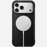 A Nomad Rugged Case for iPhone 17 Pro Max in black, with TPU bumpers, is shown from the back against a plain background. A round white MagSafe wireless charger is attached below the camera module.