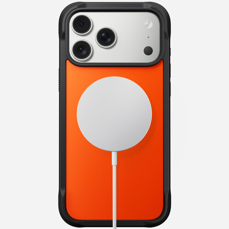 The Nomad Rugged Case - iPhone 17 Pro Max | Ultra Orange is shown from the back with TPU bumpers, MagSafe compatibility, and a round white wireless charger attached by cable. Three camera lenses and a flash are visible on the phone.