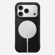 The Nomad Rugged Case for iPhone 17 Pro in black is shown from the back, displaying three rear camera lenses in the top left corner and a round white MagSafe-compatible wireless charging pad attached at the center.