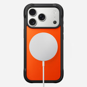 The Nomad Rugged Case - iPhone 17 Pro | Ultra Orange is shown from the back with black TPU bumpers. A white MagSafe wireless charger is attached at the center, and a charging cable extends downward.