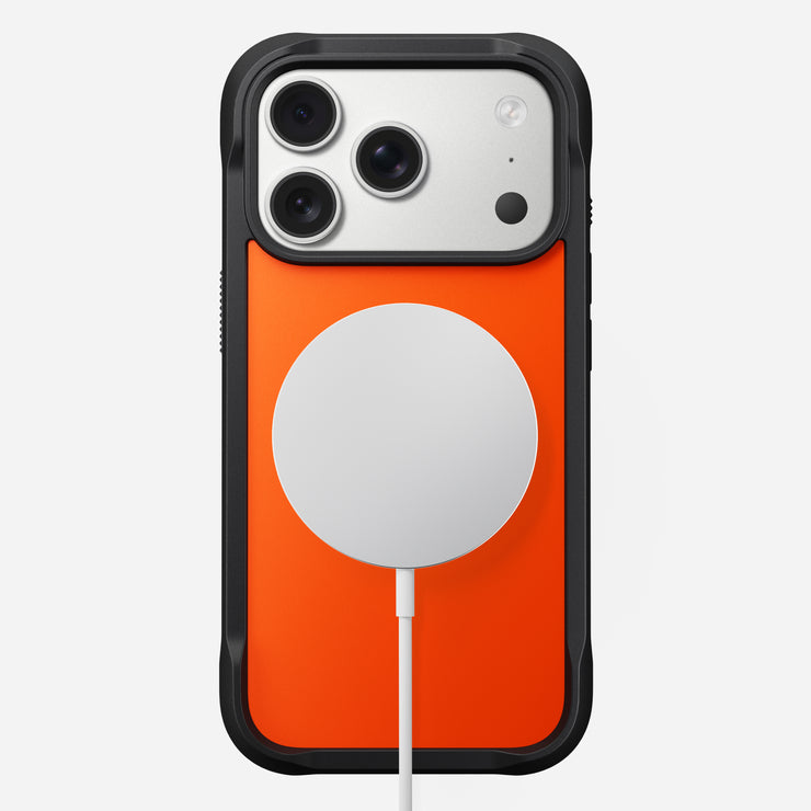 The Nomad Rugged Case - iPhone 17 Pro | Ultra Orange is shown from the back with black TPU bumpers. A white MagSafe wireless charger is attached at the center, and a charging cable extends downward.