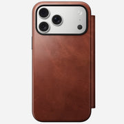 A Nomad Modern Leather Folio in Olde Dublin Horween leather covers an iPhone 17 Pro Max, showing the triple-lens camera and flash. The brown MagSafe-compatible case features a magnetic cover for easy charging.