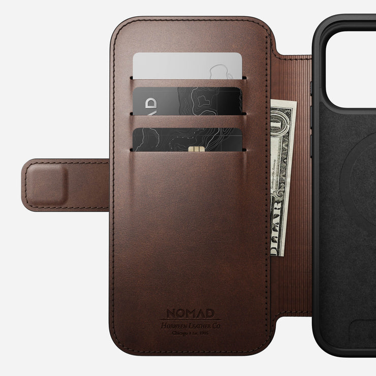 The open Nomad Modern Leather Folio for iPhone 17 Pro Max in Rustic Brown Horween leather displays three card slots holding two cards and a folded US dollar bill, along with part of the phone holder on the right.