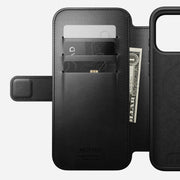 The Nomad Modern Leather Folio for iPhone 17 Pro Max in Black Horween leather is open, revealing card slots with two credit cards and a dollar bill, plus a MagSafe-compatible phone holder and an interior NOMAD Horween Leather Co. stamp.