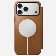 The Nomad Modern Leather Folio in English Tan-C protects the iPhone 17 Pro Max. A round white MagSafe charger attaches to the case back, and the phone features three camera lenses plus a flash in the top left corner.