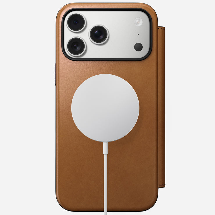 The Nomad Modern Leather Folio in English Tan-C protects the iPhone 17 Pro Max. A round white MagSafe charger attaches to the case back, and the phone features three camera lenses plus a flash in the top left corner.