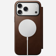 A Nomad Modern Leather Folio in brown full-grain leather encases an iPhone 17 Pro Max with a triple-lens camera, wirelessly charging on the back via a white MagSafe charger.