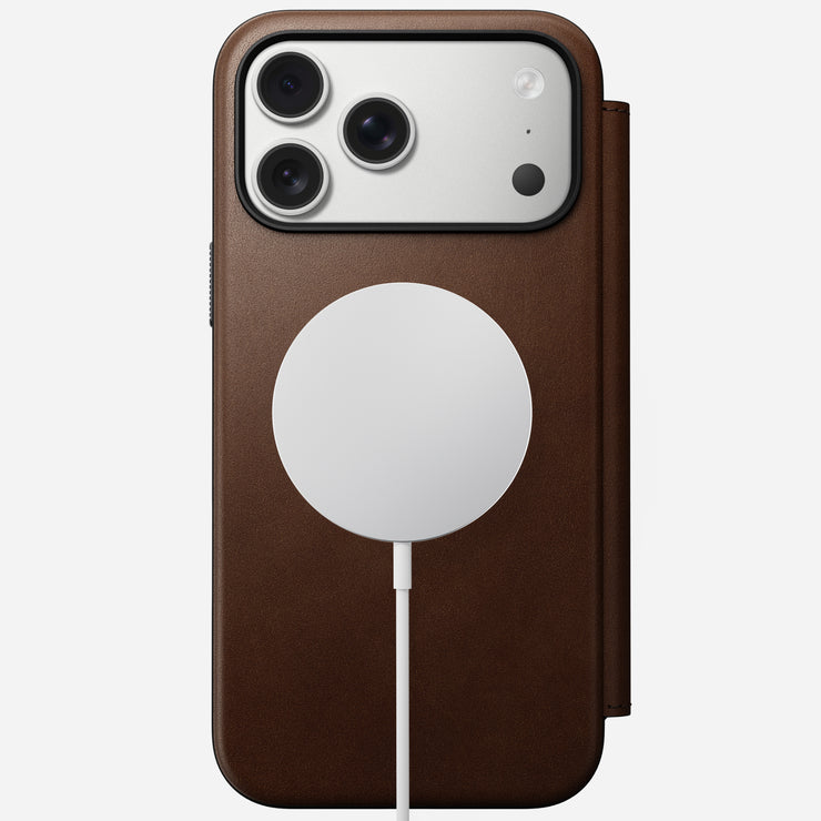 A Nomad Modern Leather Folio in brown full-grain leather encases an iPhone 17 Pro Max with a triple-lens camera, wirelessly charging on the back via a white MagSafe charger.