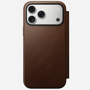 The Nomad Modern Leather Folio in brown, designed for iPhone 17 Pro Max, encases a phone with three rear cameras and a silver module, shown on a plain white background. The case is MagSafe compatible and made from premium leather.