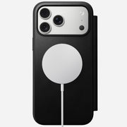 A Nomad Modern Leather Folio in black for iPhone 17 Pro Max is shown flat, viewed from above, with three camera lenses and a circular MagSafe-compatible wireless charging pad magnetically attached via a white cable.