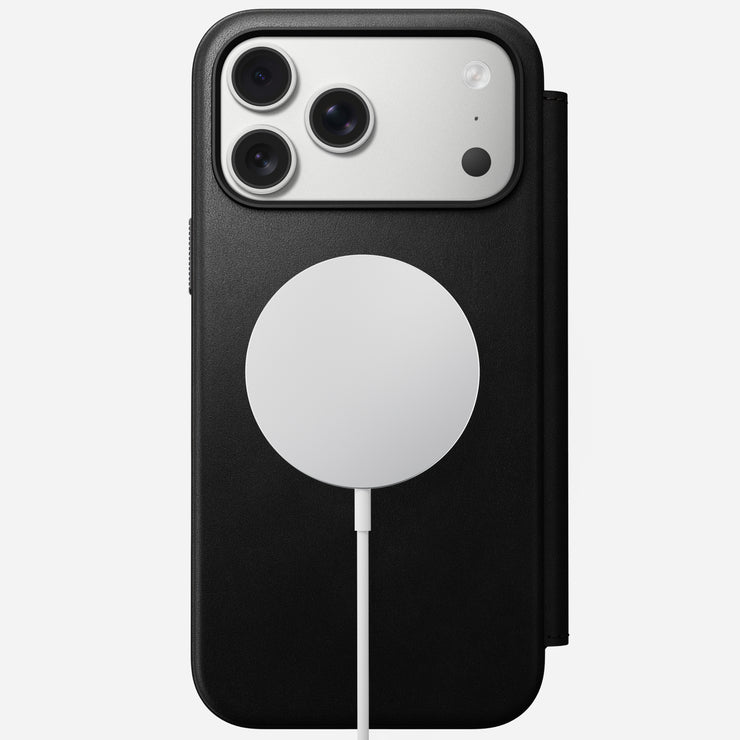 A Nomad Modern Leather Folio in black for iPhone 17 Pro Max is shown flat, viewed from above, with three camera lenses and a circular MagSafe-compatible wireless charging pad magnetically attached via a white cable.